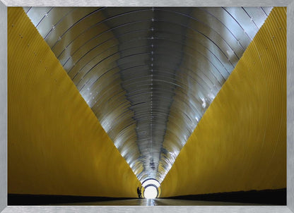 A symmetrical, perspective view down a modern, circular tunnel. The lower walls are bright yellow, creating two diagonal lines that converge towards a bright, white light at the far end. The arched ceiling is made of reflective silver metal panels, and a small silhouette stands near the exit. Artwork