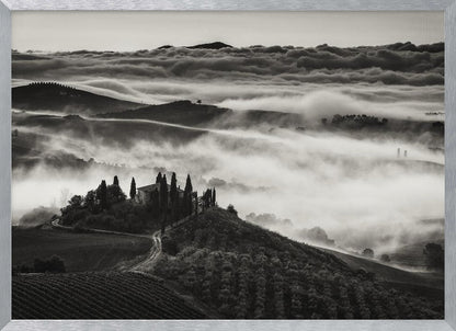 A framed, black and white photograph of a dramatic Tuscan landscape. A villa surrounded by cypress trees sits on a foreground hill, while thick fog blankets the rolling hills and valleys below. In the distance, a sea of clouds stretches towards a mountain range. Wall Art