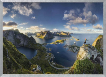 A breathtaking panoramic photograph of the Lofoten Islands in Norway from a high vantage point. A person stands on a rocky cliff with arms outstretched, overlooking a stunning landscape of deep blue fjords, lush green mountains, and the small fishing village of Reine below. The sky is a vibrant blue with scattered white clouds. Artwork