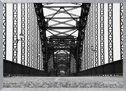 A framed, black and white, low-angle photograph capturing the symmetrical and intricate steel truss structure of a long bridge, creating a powerful vanishing point perspective down the center of the road. Poster