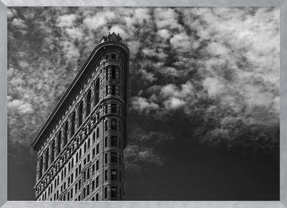 A dramatic low-angle black and white photograph of the Flatiron Building in New York City, with its sharp, triangular corner pointing towards a sky filled with fluffy, white clouds. The image is framed with a brushed silver border. Print
