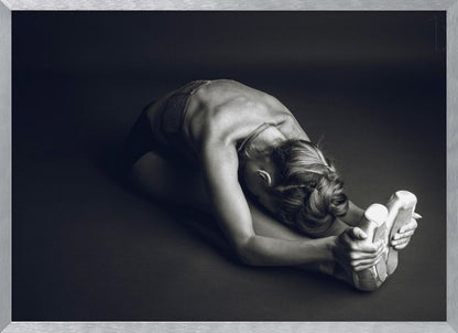 A black and white photograph of a ballerina stretching. She is seated on the floor in a deep forward fold, her torso bent over her legs as she holds onto her pointe shoes. The lighting accentuates the musculature of her back and shoulders, and her hair is styled in a braided bun. Decor