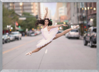 A ballerina in a white tutu and pointe shoes leaps into a split mid-air in the middle of a blurred city street, her arms extended gracefully. The image is set within a silver frame. Print