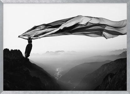 A dramatic black and white photograph of a woman standing on the edge of a mountain peak. She holds up a massive piece of dark, sheer fabric that billows in the wind, stretching across the sky. Below her, a vast, misty mountain range and a deep valley with city lights are visible. Wall Art