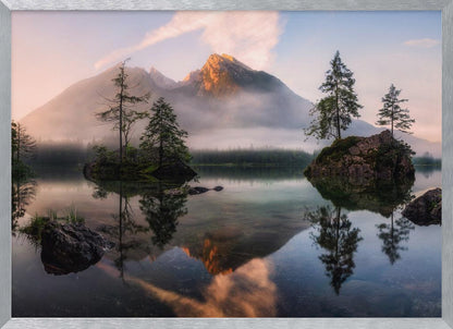 A serene mountain lake at sunrise or sunset, with a prominent peak illuminated by golden light. Small, rocky islands with pine trees dot the calm, clear water, which perfectly reflects the sky, trees, and mountains. A light mist hangs over the water and around the base of the mountains in the background. Decor