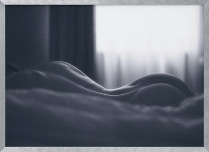 A close-up black and white artistic photograph capturing the soft curves of a person's back and hips as they lie down. The background is softly lit by a window, creating a play of light and shadow that emphasizes the human form's abstract, sculptural qualities. The image is displayed in a silver frame. Print