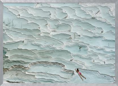 An aerial view of a woman in a pink bikini swimming in the stunning, terraced, milky-blue thermal pools of Pamukkale, Turkey. The white travertine formations create a unique, layered landscape, and the entire image is enclosed in a silver frame. Artwork