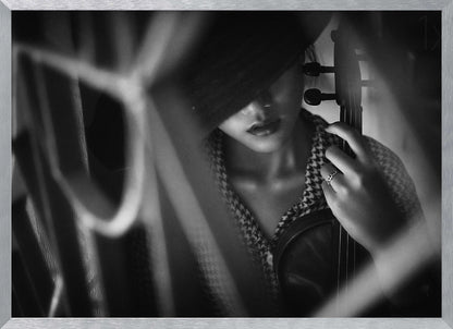A moody, artistic black and white photograph of a woman holding a violin. Her face is partially obscured by a dark hat, creating a mysterious atmosphere. The image is shot through a blurred foreground, adding depth and intimacy to the portrait. Poster