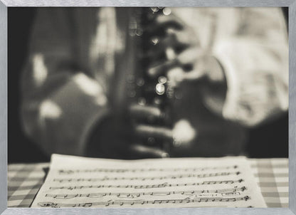 A shallow depth of field, black and white photograph focusing on sheet music in the foreground. The background is softly blurred, showing the hands of a musician holding a woodwind instrument. Wall Art