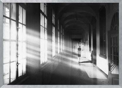 A black and white photograph of a woman walking away down a long, dark hallway. Bright shafts of sunlight pour through large windows on the left, creating a dramatic interplay of light and shadow on the floor and walls. Poster