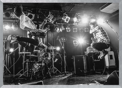 A high-energy, black and white photograph of a rock band performing live. The bassist and guitarist are frozen in mid-air as they jump, while the drummer plays in the background, all under bright stage lights. Print