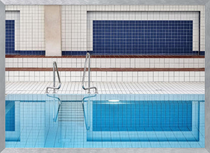 A framed, symmetrical photograph of a minimalist indoor swimming pool with blue and white tiles, stainless steel ladders, and a calm, reflective water surface. Poster