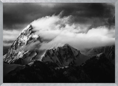 A dramatic black and white photograph of a snow-covered mountain peak being enveloped by thick, swirling clouds under a dark, stormy sky. The rugged slopes and sharp ridges of the mountain are highlighted by the snow, while a dark forest sits at the base. The entire image is presented within a textured silver frame. Print