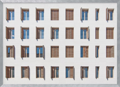 A symmetrical architectural photo of a modern white building facade featuring a grid of twenty-eight windows with wooden louvered shutters, some open and some closed, creating a varied pattern, all within a brushed silver frame. Decor
