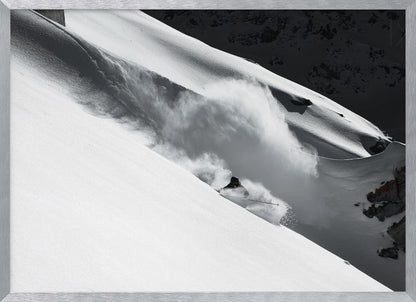 A dramatic black and white photograph of a skier speeding down a steep, snow-covered mountain, kicking up a large cloud of powder. The image is framed with a brushed silver border. Decor
