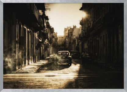 A sepia-toned, atmospheric photograph of a classic car driving down a narrow street in an old city. The sun hangs low, casting long shadows and illuminating smoke or dust in the golden air. Artwork