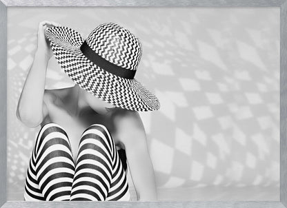 A black and white artistic photograph of a woman sitting down, her face obscured by a large, wide-brimmed sun hat with a black and white chevron pattern. She is wearing black and white striped leggings and holding the brim of her hat with one hand. The image is set against a plain background with patterned shadows and is presented within a silver frame. Artwork
