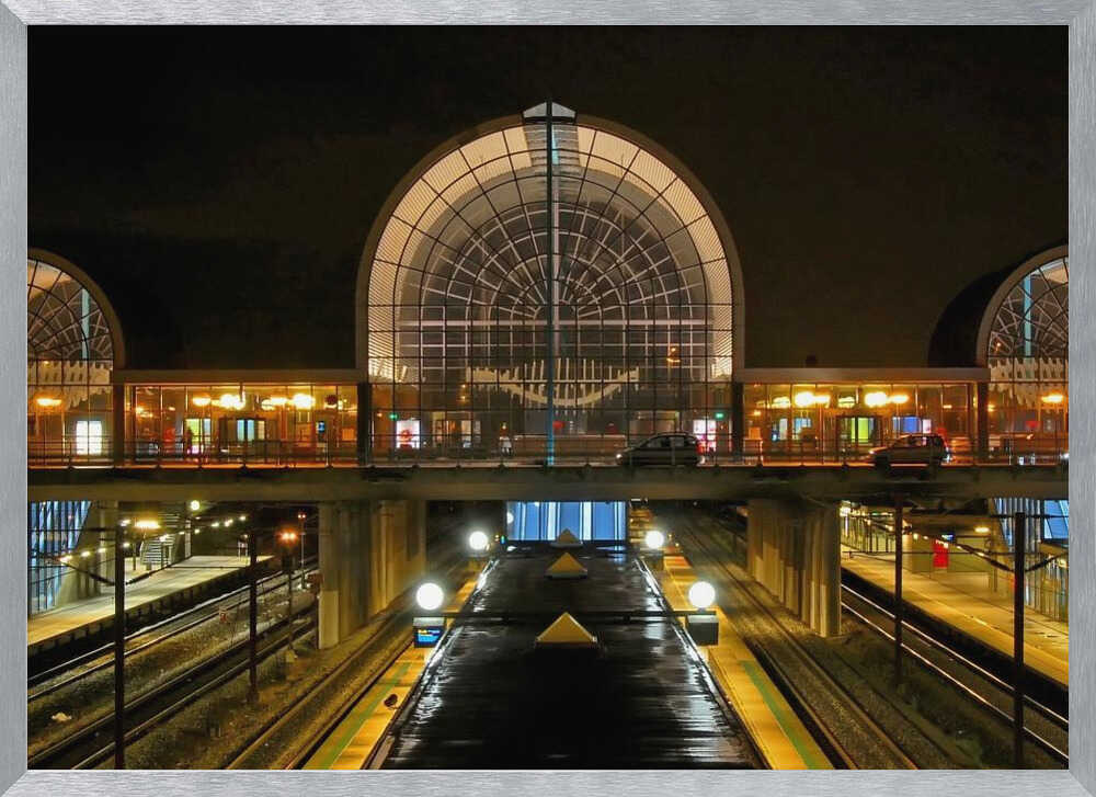 A symmetrical view of a modern train station at night, with a large illuminated arched glass window. Below, empty, wet train tracks and platforms reflect the station's lights, while cars cross an overpass in the middle ground. Wall Art