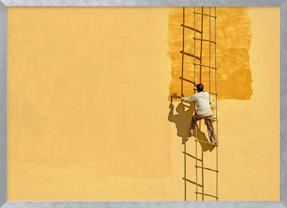 A man viewed from behind stands on a rope ladder, painting a large, bright yellow wall a slightly darker shade of yellow, casting a stark shadow in the bright sun. The image is presented within a silver frame. Decor