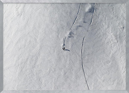 An aerial, top-down photograph of a lone snowboarder carving a turn down a vast, textured, snowy mountain slope, leaving fresh tracks in the powder. The image is presented in a simple, grey frame. Wall Art