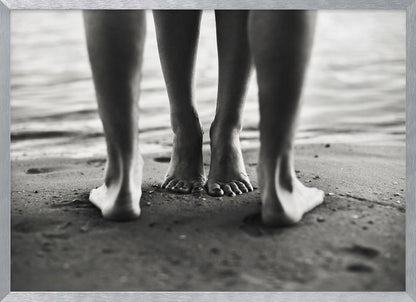 A framed, black and white photograph showing a low-angle view of two people's bare feet and lower legs standing on a wet sandy beach. The central pair of feet is in focus, while another person's legs in the foreground are blurred, creating depth. The water's edge is visible in the background. Artwork