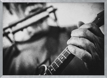A grainy, black and white close-up photograph of a musician's hand gripping the neck of a stringed instrument. The focus is on the hand and the frets, with the musician's body and face softly blurred in the background. The image is enclosed in a silver frame. Wall Art