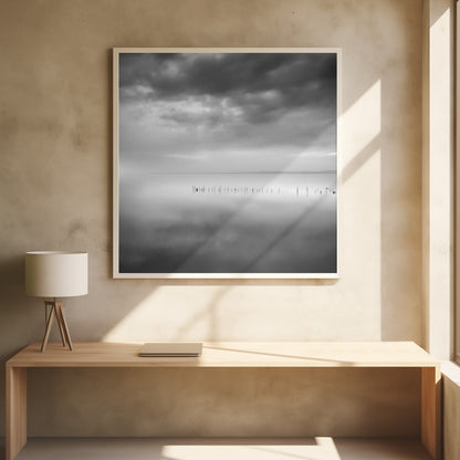 A minimalist black and white photograph in a light wood frame, depicting a calm, serene lake under a heavy, cloudy sky. The still water reflects the sky, with a row of weathered wooden posts breaking the surface along the horizon. Print