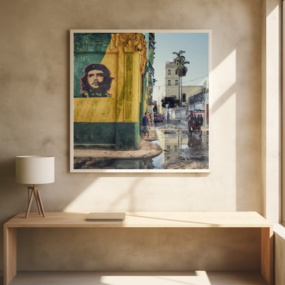 A framed photograph of a vibrant street corner in a city like Havana, Cuba, with a large mural of Che Guevara on a yellow and green wall. The wet street reflects the buildings and a palm tree, while people, a vintage car, and a rickshaw occupy the narrow road. Poster