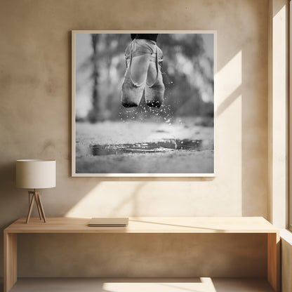 A close-up, black and white photograph of a ballerina's feet in worn pointe shoes, captured in mid-air above a puddle, with water splashing upwards. The motion is frozen, highlighting the contrast between grace and a raw outdoor element. The image is presented in a light wood frame. Poster