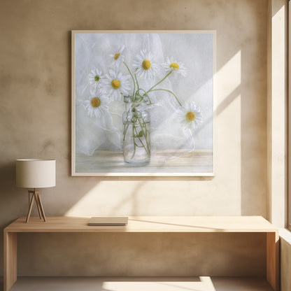 A delicate still life photograph of several white daisies with bright yellow centers in a clear glass jar. The arrangement sits on a light wooden surface against a backdrop of soft, draped white fabric. The entire image is enclosed in a light wood frame. Artwork