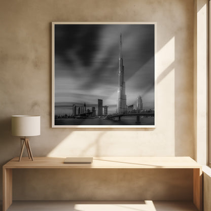 A framed, long-exposure black and white photograph of the Dubai skyline, featuring the iconic Burj Khalifa. The sky has a dramatic, streaky appearance from the long exposure, contrasting with the sharp details of the modern architecture. A bridge crosses the water in the foreground. Print