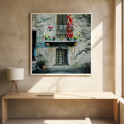A woman in a vibrant red dress stands on an ornate white balcony filled with colorful potted flowers. The building has a weathered, gray stucco facade. To the left, a small child in a blue dress peeks out from a shadowed doorway, adding a layer of story to the rustic scene. Decor