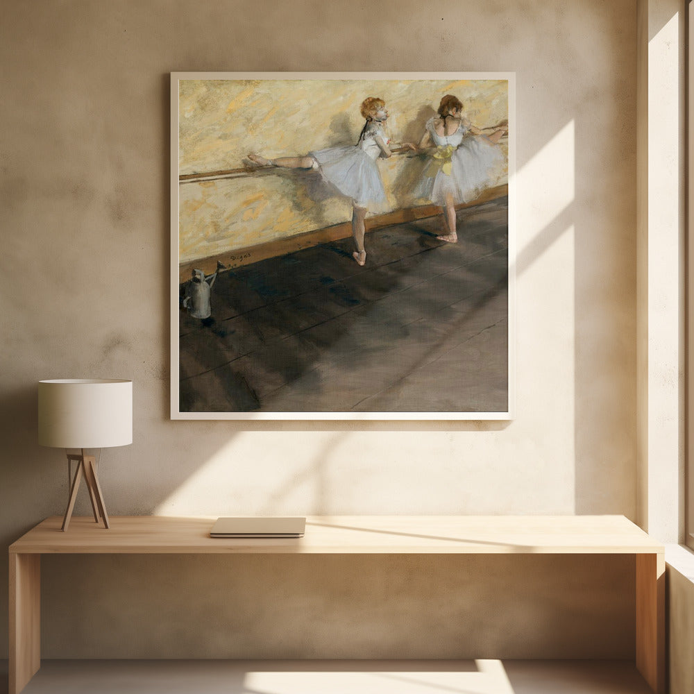 A framed impressionist painting by Edgar Degas depicting two ballet dancers in white tutus practicing at a barre. The studio has a yellow wall and a dark brown wooden floor. One dancer stretches her leg on the barre while the other stands beside her. Artwork