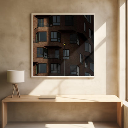 A close-up photograph of a modern red-brick apartment building, framed in light wood. A sharp shadow line divides the facade, with a lit streetlamp providing a warm yellow glow in the shaded area. Decor