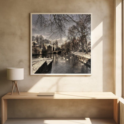 A vintage-style, framed photograph of a European canal scene in winter. Historic buildings and bare trees line the partially frozen canal, where several white swans are swimming. A light dusting of snow covers the canal banks and rooftops under a cloudy sky. Wall Art
