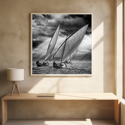 A dramatic black and white photograph of a fleet of traditional wooden sailboats with large, billowing sails racing on a choppy sea under a dark, stormy sky. The image is presented in a light wood frame. Print