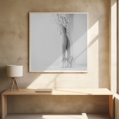 A framed, grainy black and white photograph of a dancer's legs and feet. The dancer stands on tiptoes against a white wall, wearing a delicate lace skirt. Poster