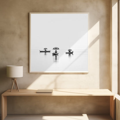 A minimalist black and white photograph of three fishermen in small boats on a calm, misty lake, each holding an umbrella. The image is framed in light wood. Artwork