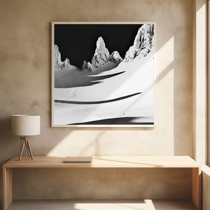 A framed, high-contrast black and white photograph of a majestic, snow-covered mountain range. Jagged peaks are sunlit against a stark black sky, casting long, dramatic shadows across a vast, pristine snowfield below. A tiny, solitary figure stands in the snow, highlighting the immense scale of the landscape. Print